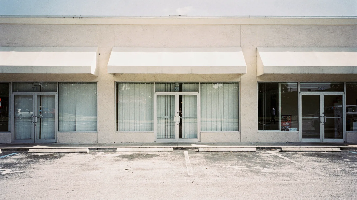 A row of Southwest Florida strip mall storefronts in flat bright midday sun, the units on either side open while the middle one sits dark with its blinds drawn.