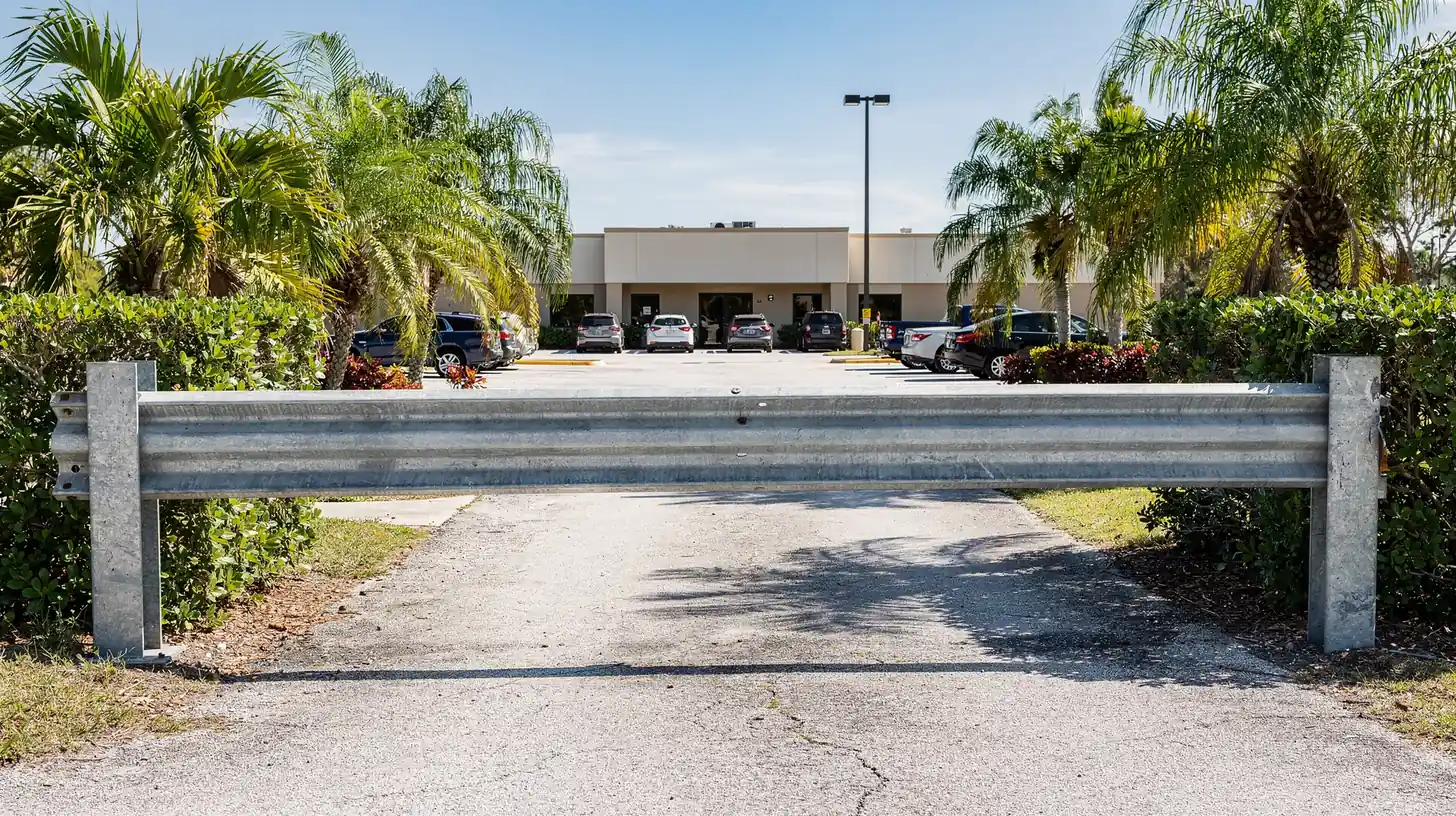 A metal barrier gate lowered across the entrance to a small Florida commercial driveway in bright morning sun, the building and parking lot open and visible beyond it.