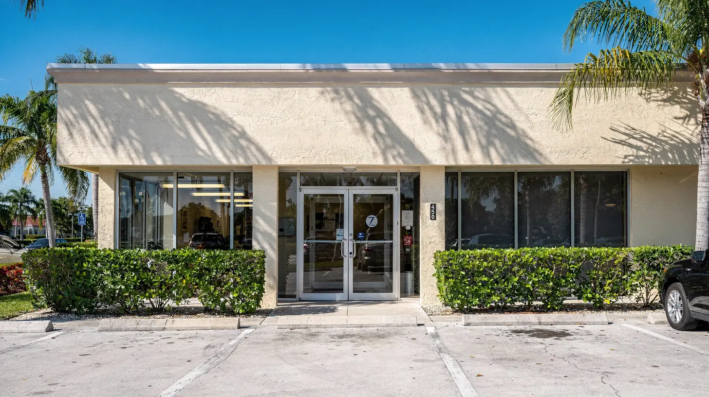 The glass entrance and pale stucco frontage of a small Florida dental practice in bright mid morning sun, hard palm shadows falling across the wall.
