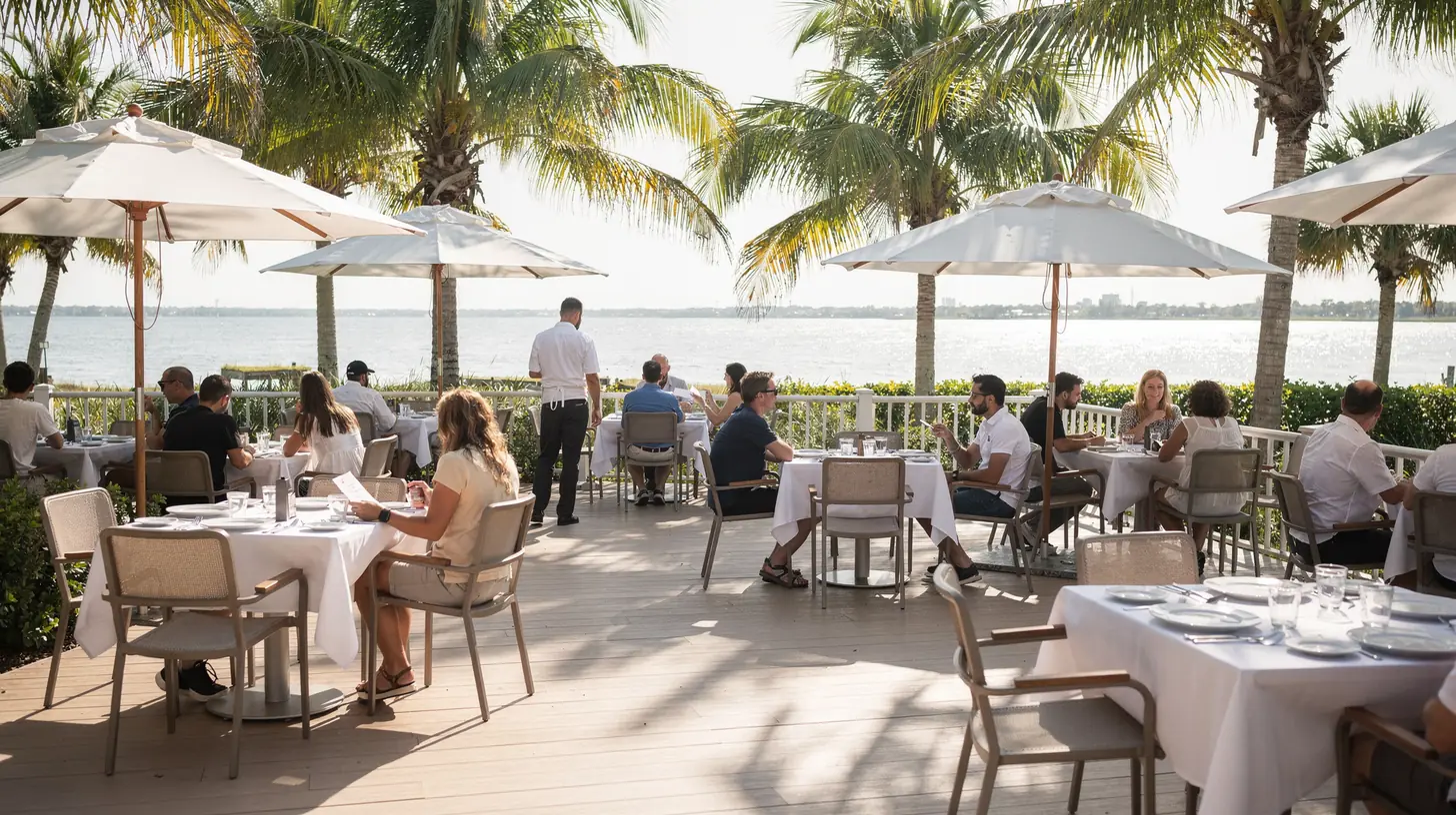 A Gulf coast Florida restaurant patio during lunch service, white umbrellas and set tables under dappled palm shade with calm water beyond.