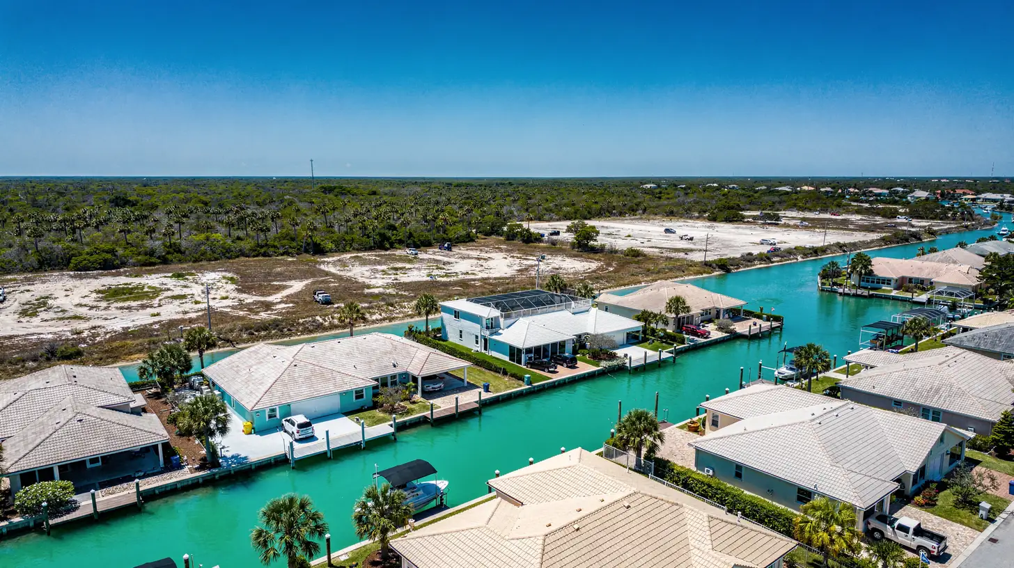 A bright aerial view over a Gulf coast Florida canal neighborhood of pale tile roofs and turquoise canals meeting undeveloped palmetto scrub.
